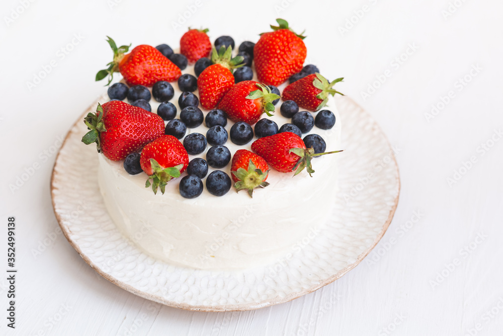 A beautiful appetizing cake with white cream frosting and fresh summer berries on hand made plate on white wooden background. Delicious home made dessert with blueberries and strawberries A beautiful appetizing cake with white cream frosting and fresh summer berries on hand made plate on white wooden background. Delicious home made dessert with blueberries and strawberries