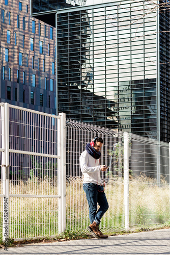 Young bearded male with headphones and holding smartphone while leaning on a metallic fence against skyscrapers in sunny day