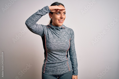 Young blonde fitness woman wearing sport workout clothes over isolated background very happy and smiling looking far away with hand over head. Searching concept.