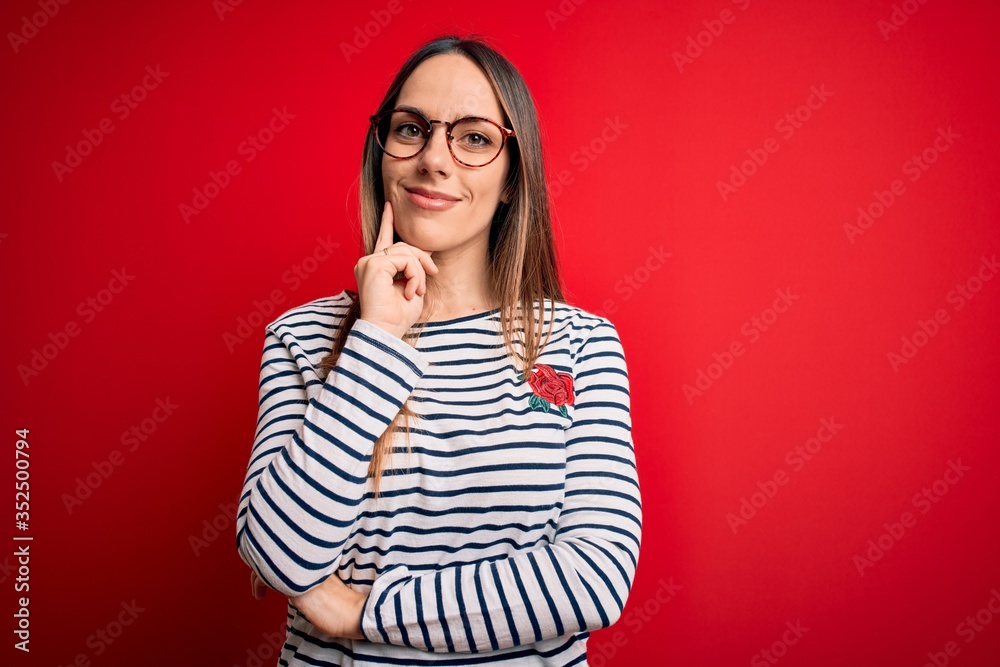 Young beautiful blonde woman with blue eyes wearing glasses standing over red background looking confident at the camera smiling with crossed arms and hand raised on chin. Thinking positive. Young beautiful blonde woman with blue eyes wearing glasses standing over red background looking confident at the camera smiling with crossed arms and hand raised on chin. Thinking positive.