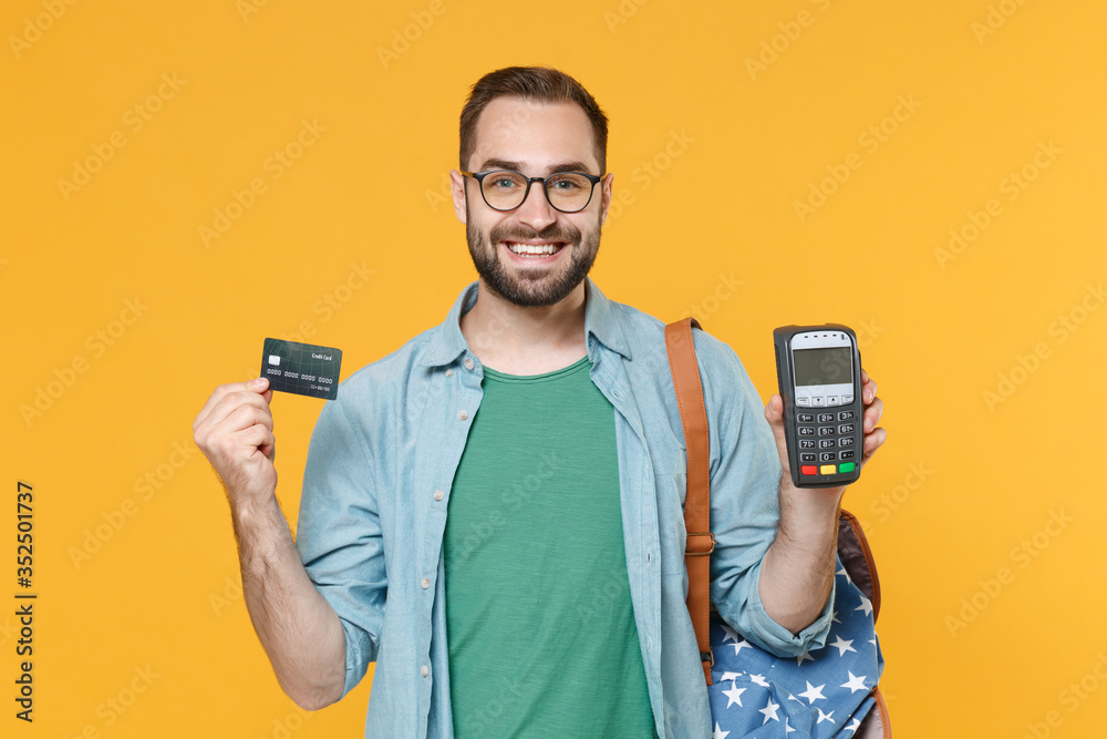Smiling young man student in glasses backpack isolated on yellow background. Education in high school university college concept. Hold bank payment terminal to process acquire credit card payments. Smiling young man student in glasses backpack isolated on yellow background. Education in high school university college concept. Hold bank payment terminal to process acquire credit card payments.