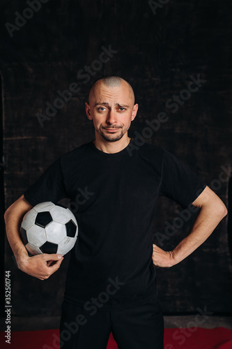 A male sports teacher stands against a dark background, holding a soccer ball