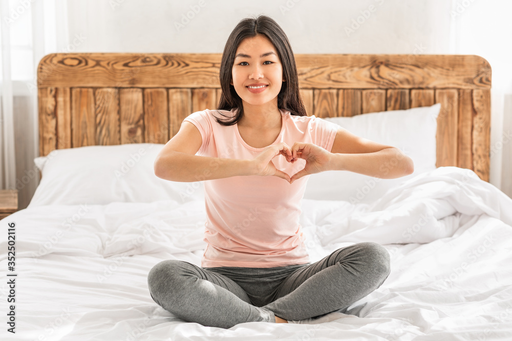 Asian Woman Gesturing Heart Shape Sitting On Bed At Home Asian Woman Gesturing Heart Shape Sitting On Bed At Home
