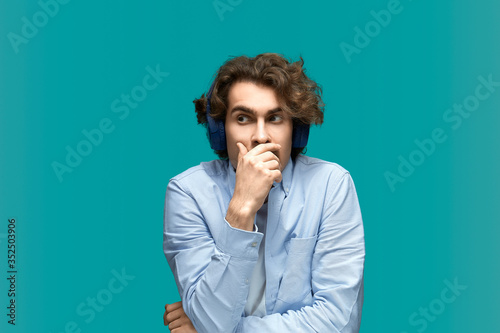 May not notice. Portrait of a young beautiful man wearing white t-shirt and blue shirt in headphones closes his mouth with his hands because he blurted something out