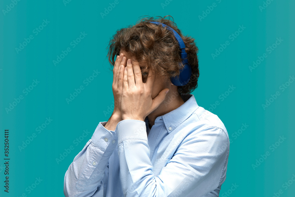 Cheater. Portrait of a young beautiful man wearing white t-shirt and blue shirt in headphones covering face with palms and peeking through fingers with one eye