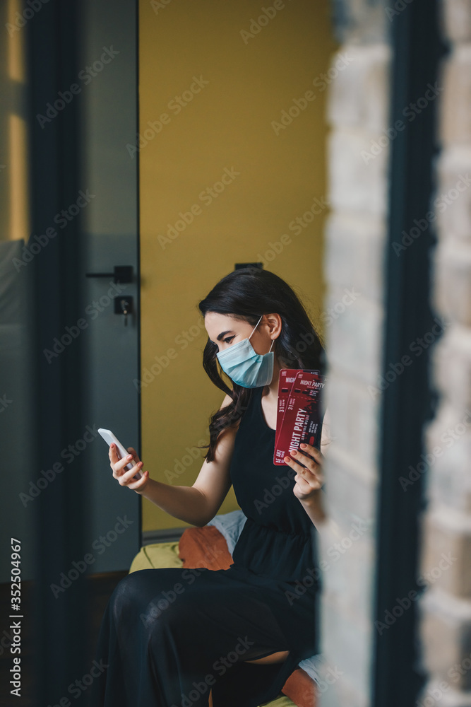 selective focus of young woman in medical mask and black dress holding night party tickets and using smartphone, end of quarantine concept selective focus of young woman in medical mask and black dress holding night party tickets and using smartphone, end of quarantine concept