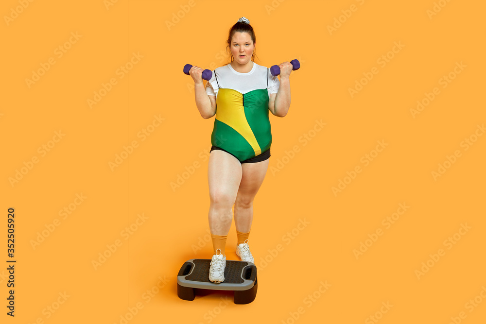 Fat confident athlete with dumbbells in her hands is training on the step platform Fat confident athlete with dumbbells in her hands is training on the step platform
