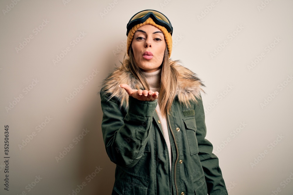 Young brunette skier woman wearing snow clothes and ski goggles over white background looking at the camera blowing a kiss with hand on air being lovely and sexy. Love expression. Young brunette skier woman wearing snow clothes and ski goggles over white background looking at the camera blowing a kiss with hand on air being lovely and sexy. Love expression.