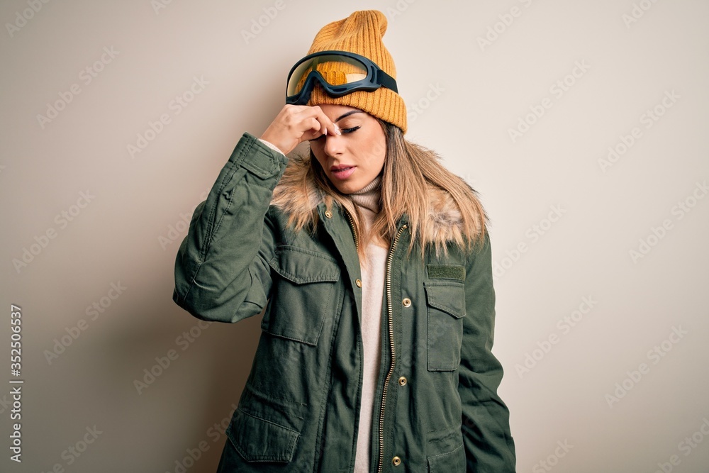 Young brunette skier woman wearing snow clothes and ski goggles over white background tired rubbing nose and eyes feeling fatigue and headache. Stress and frustration concept. Young brunette skier woman wearing snow clothes and ski goggles over white background tired rubbing nose and eyes feeling fatigue and headache. Stress and frustration concept.