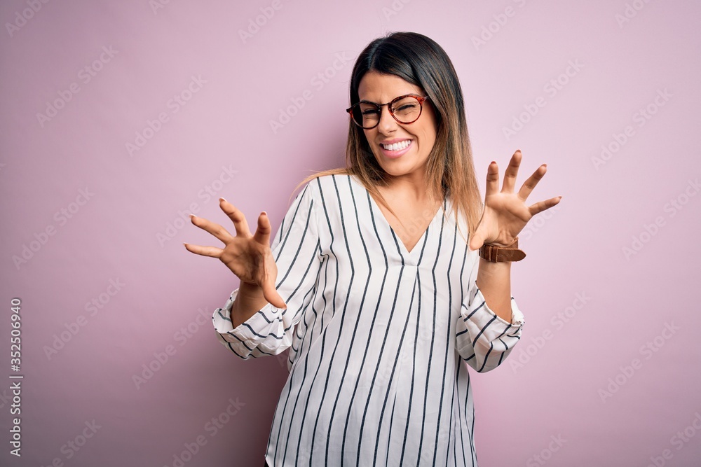 Young beautiful woman wearing casual striped t-shirt and glasses over pink background smiling funny doing claw gesture as cat, aggressive and sexy expression Young beautiful woman wearing casual striped t-shirt and glasses over pink background smiling funny doing claw gesture as cat, aggressive and sexy expression