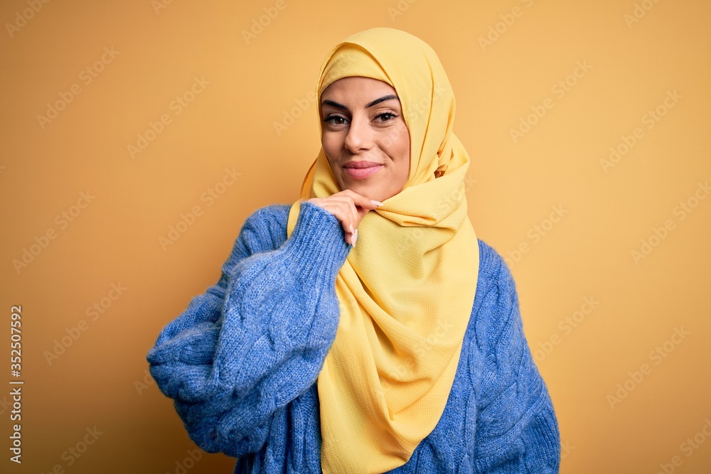 Young beautiful brunette muslim woman wearing arab hijab over isolated yellow background looking confident at the camera smiling with crossed arms and hand raised on chin. Thinking positive. Young beautiful brunette muslim woman wearing arab hijab over isolated yellow background looking confident at the camera smiling with crossed arms and hand raised on chin. Thinking positive.
