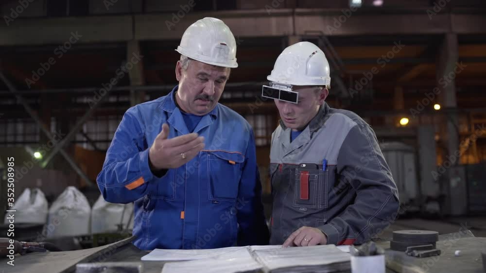 Two workers at heavy industry factory have conversation while looking at blueprint. Engineers explore and discuss project in heavy industry factory. Foundry for Metal Cast and Production. Two workers at heavy industry factory have conversation while looking at blueprint. Engineers explore and discuss project in heavy industry factory. Foundry for Metal Cast and Production.