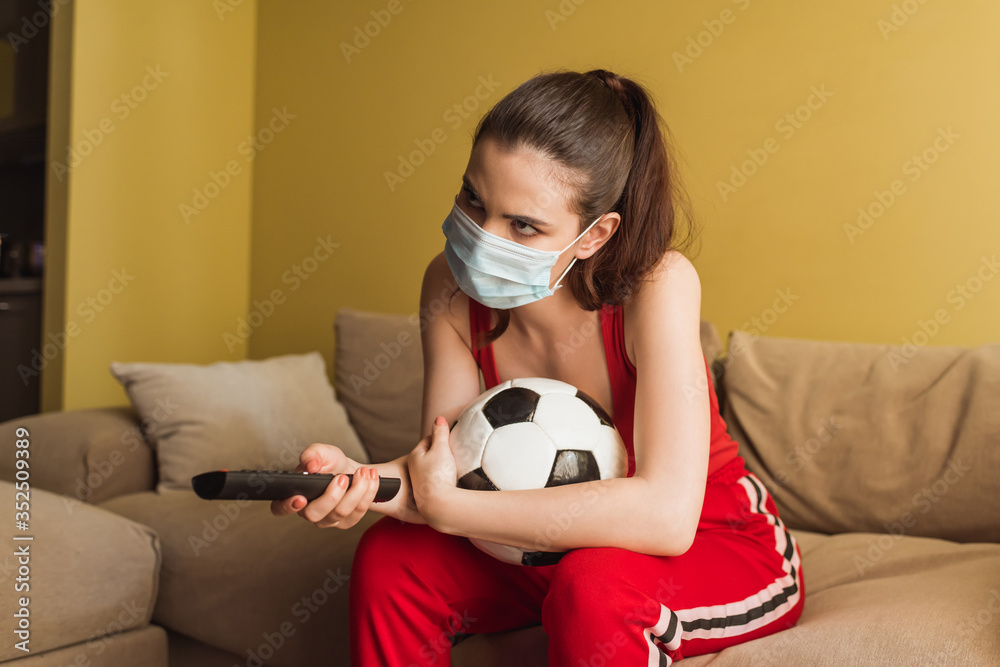 sportive girl in medical mask holding remote controller near football while watching championship in living room sportive girl in medical mask holding remote controller near football while watching championship in living room