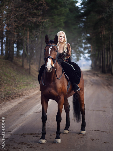 Beautiful girl dressed in stylish medieval clothes is training a beautiful horse in the forest. Fashion and style concept.