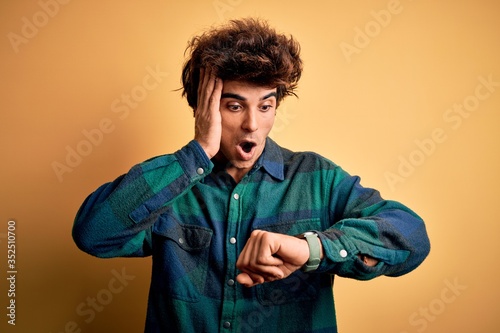Young handsome man wearing casual shirt standing over isolated yellow background Looking at the watch time worried, afraid of getting late