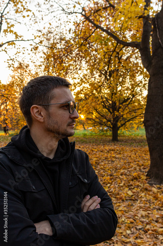 A young short-haired guy wearing glasses and a black jacket stands with his arms crossed in the autumn park. Meditation in the falling park.