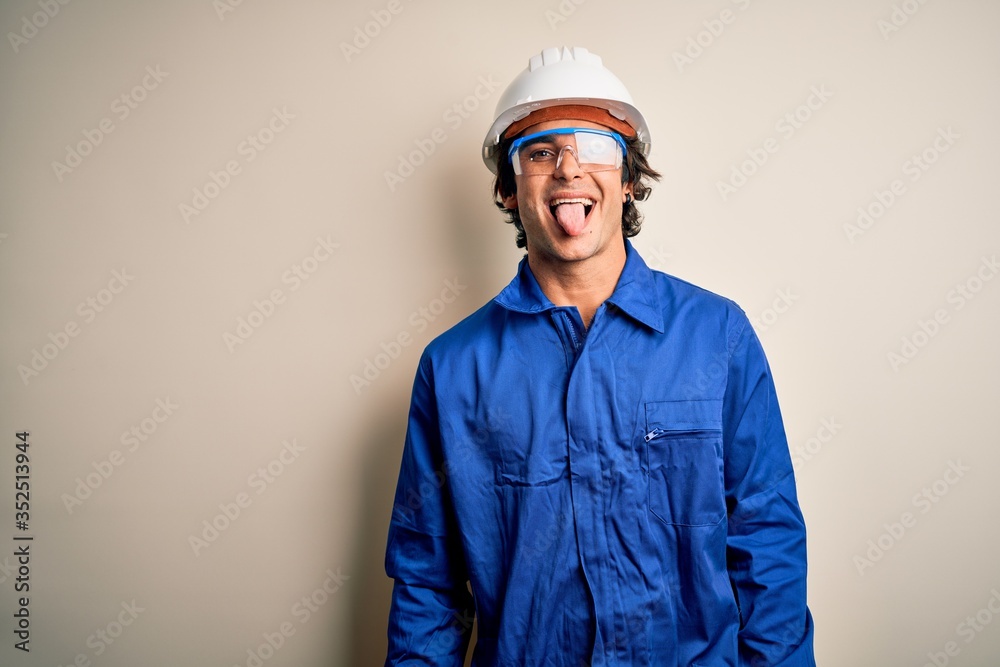 Young constructor man wearing uniform and security helmet over isolated white background sticking tongue out happy with funny expression. Emotion concept. Young constructor man wearing uniform and security helmet over isolated white background sticking tongue out happy with funny expression. Emotion concept.