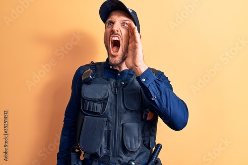 Young handsome policeman wearing police uniform and bulletprof over yellow background shouting and screaming loud to side with hand on mouth. Communication concept.