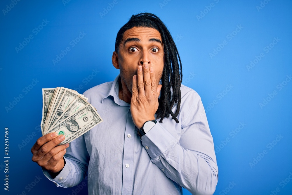 Young african american afro man with dreadlocks holding dollars over blue background cover mouth with hand shocked with shame for mistake, expression of fear, scared in silence, secret concept Young african american afro man with dreadlocks holding dollars over blue background cover mouth with hand shocked with shame for mistake, expression of fear, scared in silence, secret concept