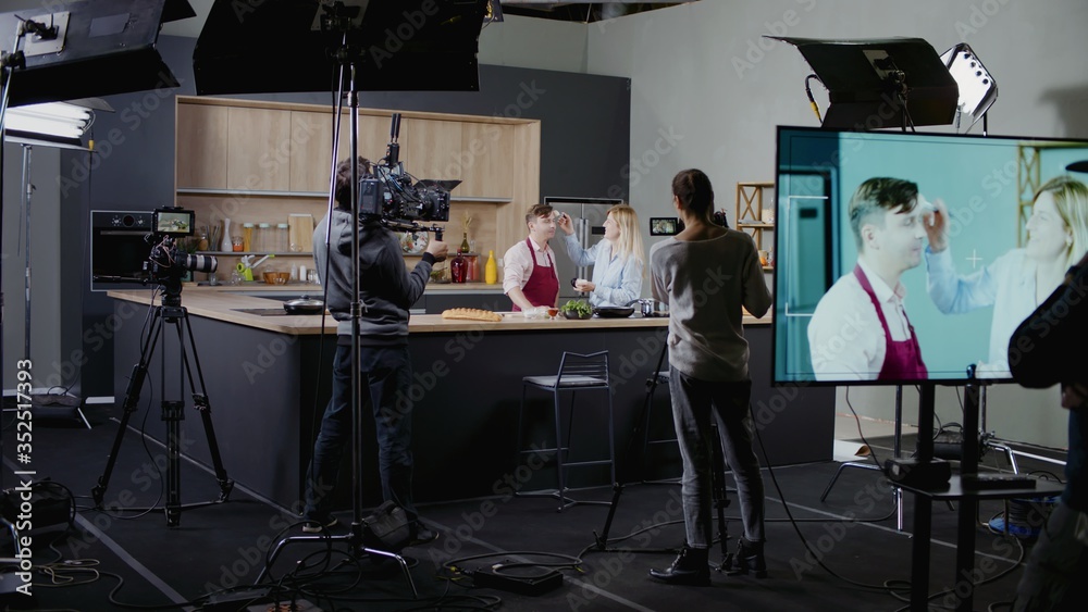 WIDE Behind the scenes of studio set, makeup artist make corrections during shooting television cooking show featuring celebrity chef, professional TV production