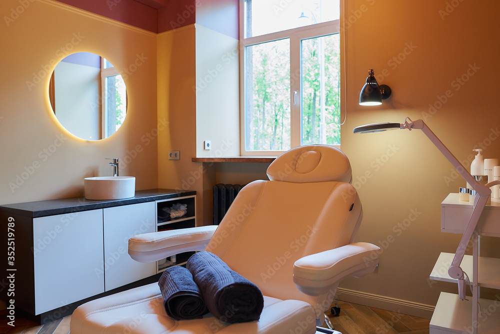 Interior of the cosmetology office with a sink, a round mirror, an electrical facial beauty bed and chair, a trolley cart with skincare products, a led lamp in a beauty salon. A window curtain raised.