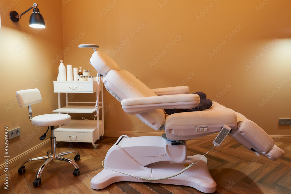 Modern interior of the cosmetology office with a full-electrical facial beauty bed and chair, a salon stool, a trolley cart with skincare products, and a led lamp in a beauty salon.