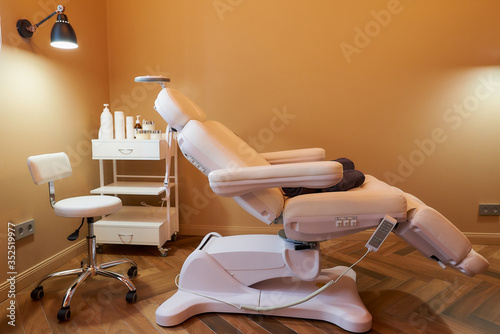 Obraz na płótnie Modern interior of the cosmetology office with a full-electrical facial beauty bed and chair, a salon stool, a trolley cart with skincare products, and a led lamp in a beauty salon. w salonie