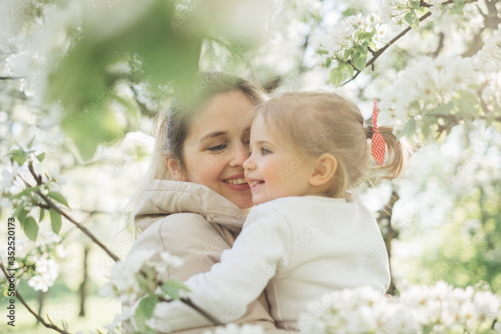 A beautiful charming mother and daughter walk in a flowering park in spring. Warm hugs, tenderness, kiss mom and baby. Blooming white trees, the sun, good mood, joy. A beautiful charming mother and daughter walk in a flowering park in spring. Warm hugs, tenderness, kiss mom and baby. Blooming white trees, the sun, good mood, joy.