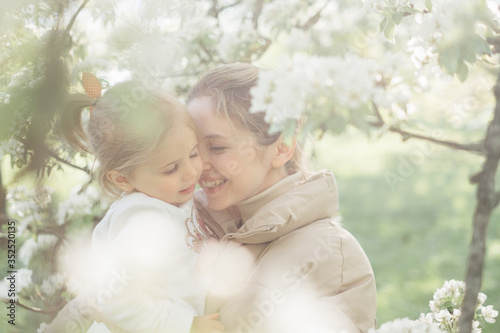 A beautiful charming mother and daughter walk in a flowering park in spring. Warm hugs, tenderness, kiss mom and baby. Blooming white trees, the sun, good mood, joy.