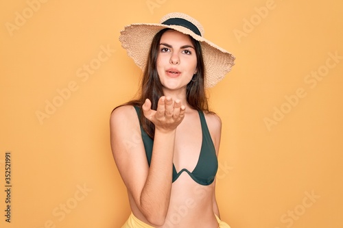 Young beautiful girl wearing swimwear bikini and summer sun hat over yellow background looking at the camera blowing a kiss with hand on air being lovely and sexy. Love expression.