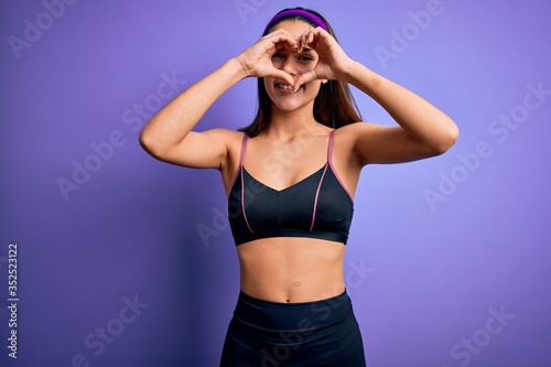 Young beautiful sporty girl doing sport wearing sportswear over isolated purple background Doing heart shape with hand and fingers smiling looking through sign © Krakenimages.com