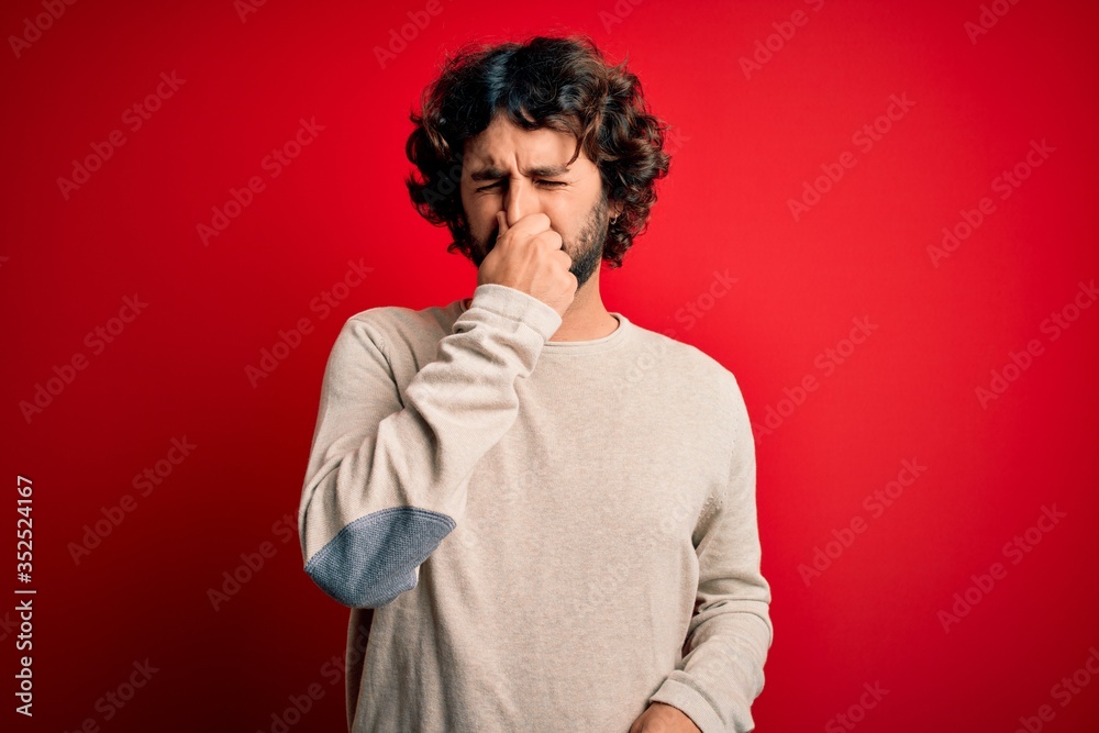 Young handsome man with beard wearing casual sweater standing over red background smelling something stinky and disgusting, intolerable smell, holding breath with fingers on nose. Bad smell