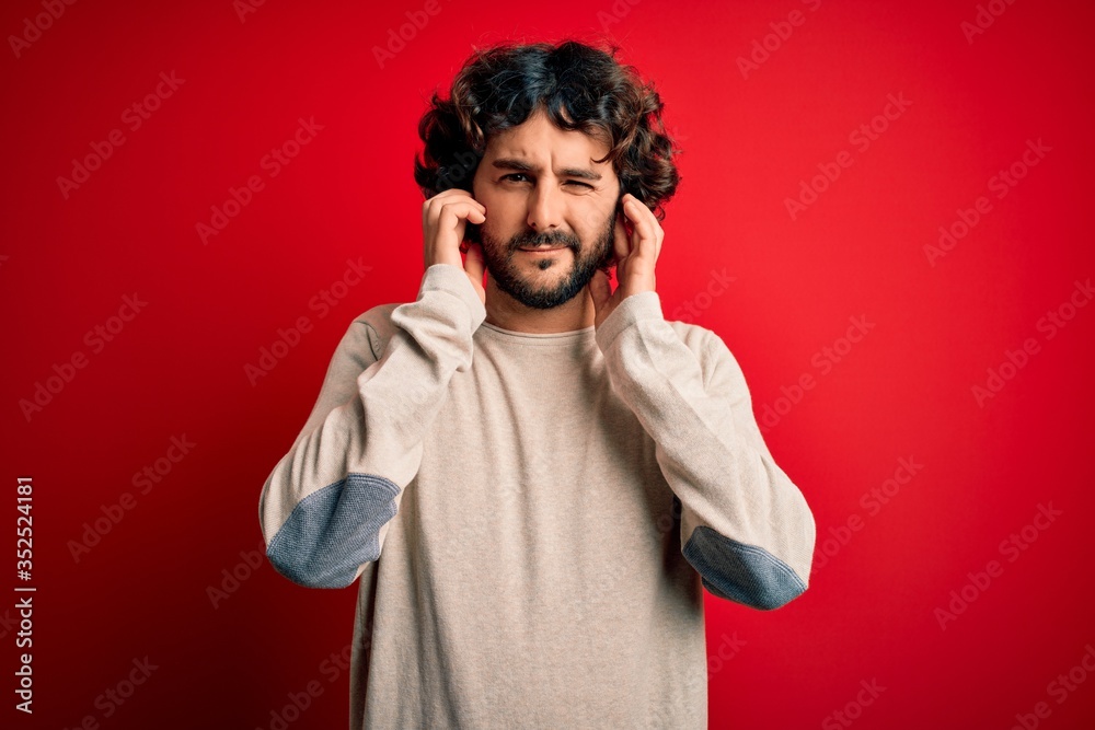 Young handsome man with beard wearing casual sweater standing over red background covering ears with fingers with annoyed expression for the noise of loud music. Deaf concept. Young handsome man with beard wearing casual sweater standing over red background covering ears with fingers with annoyed expression for the noise of loud music. Deaf concept.