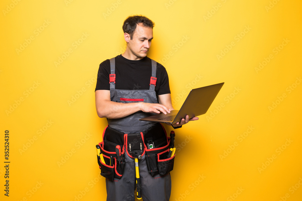 Young man worker with tools belt with laptop isolated on yellow background Young man worker with tools belt with laptop isolated on yellow background