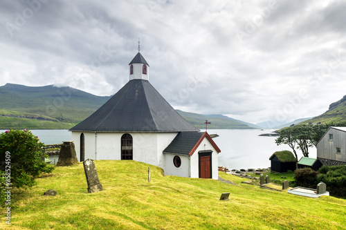 Summer view of traditional church in faroese village. Beauty landscape with foggy fjord and high mountains. Faroe Islands, Denmark.