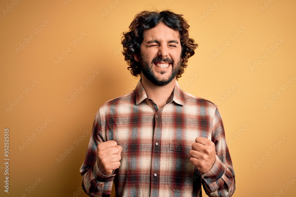 Obraz premium Young handsome man with beard wearing casual shirt standing over yellow background excited for success with arms raised and eyes closed celebrating victory smiling. Winner concept.