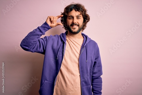 Young handsome sporty man with beard wearing casual sweatshirt over pink background smiling and confident gesturing with hand doing small size sign with fingers looking and the camera. Measure