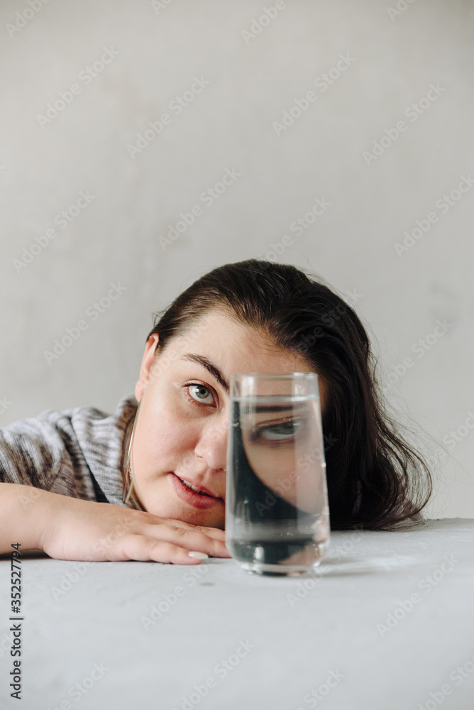 Fototapeta premium beautiful girl at the table on a gray background in her hands with a glass of water. Girl's face through a glass of water