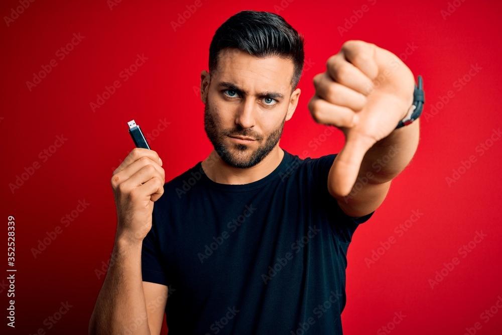 Young man holding USB memory data drive over red isolated background with angry face, negative sign showing dislike with thumbs down, rejection concept Young man holding USB memory data drive over red isolated background with angry face, negative sign showing dislike with thumbs down, rejection concept