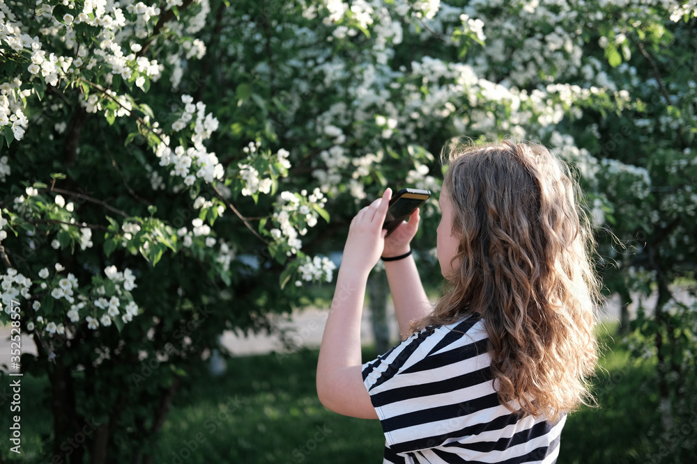 beautiful curly girl taking picture of blooming tree on a smartphone. teenager girl before a bush with white flowers. walking outdoors after quarantine. calm and peaceful place. 