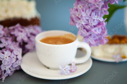 Fresh espresso in a white cup and lilac flowers on the background.
