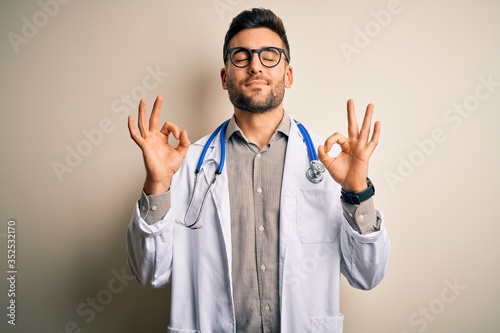 Young doctor man wearing glasses, medical white robe and stethoscope over isolated background relax and smiling with eyes closed doing meditation gesture with fingers. Yoga concept. © Krakenimages.com