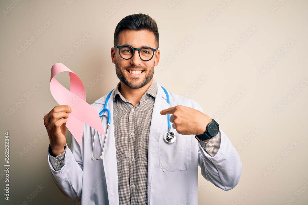 Young doctor man wearing stethoscope holding pink ribbon about cancer over isolated background with surprise face pointing finger to himself Young doctor man wearing stethoscope holding pink ribbon about cancer over isolated background with surprise face pointing finger to himself