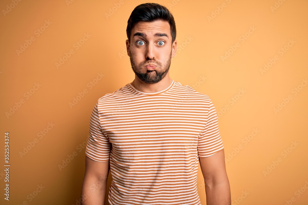 Young handsome man with beard wearing casual striped t-shirt over yellow background puffing cheeks with funny face. Mouth inflated with air, crazy expression. Young handsome man with beard wearing casual striped t-shirt over yellow background puffing cheeks with funny face. Mouth inflated with air, crazy expression.