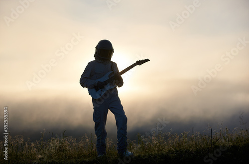 Silhouette of space traveler playing melody on guitar in misty grassy valley with white mystical sky on background. Astronaut in while space suit and helmet. Concept of music, astronautics and nature.