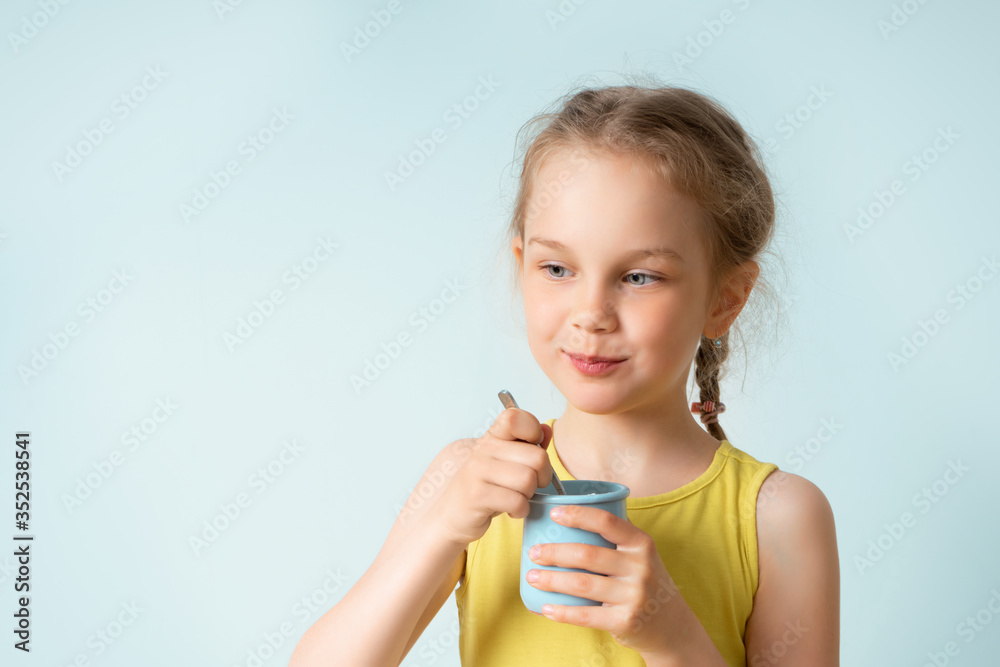 Cute little girl with funny pigtails holds plastic cup with yoghurt and a teaspoon preparing to eat. Happy childhood, emotions, gesturing, healthy food. Close up studio portrait isolated on light blue Cute little girl with funny pigtails holds plastic cup with yoghurt and a teaspoon preparing to eat. Happy childhood, emotions, gesturing, healthy food. Close up studio portrait isolated on light blue
