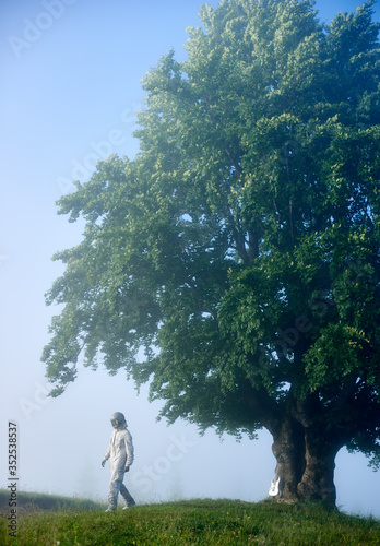 Spaceman in space suit walking away from tree in foggy morning leaving his guitar under the big old green tree.