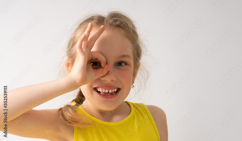 Naughty little girl with adorable smile looking at camera through her fingers showing okay gesture. Happy childhood, emotions, gesturing, sly, having fun. Close up studio portrait isolated on white Naughty little girl with adorable smile looking at camera through her fingers showing okay gesture. Happy childhood, emotions, gesturing, sly, having fun. Close up studio portrait isolated on white