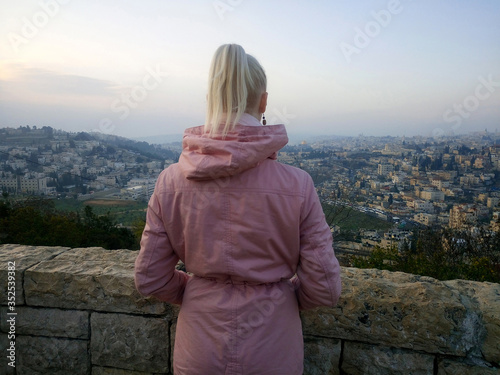 The blonde girl is looking at Jerusalem. View of a girl from the back in a pink jacket.