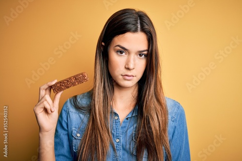 Young beautiful girl holding healthy protein bar standing over isolated yellow background with a confident expression on smart face thinking serious